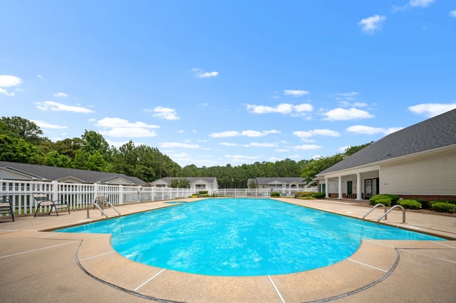 Bright blue swimming pool with lounge chairs and white fence surrounding it, with apartment buildings in the background at Ashton Pointe apartments in Monroe, GA.