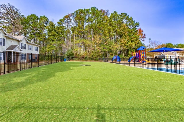 Fenced-in dog park with artificial turf and playground in the background.