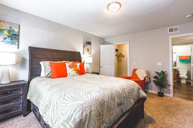 King-size bed with orange and floral pillows in a bedroom with a closet and ensuite bathroom. Explore apartments for rent in East Point GA near Atlanta airport—schedule your tour today or call for more information.
