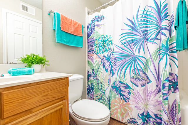 Bathroom with a wooden vanity, white toilet, and a tropical-themed shower curtain View apartments for rent in East Point GA with parking—schedule your tour or call for more information.