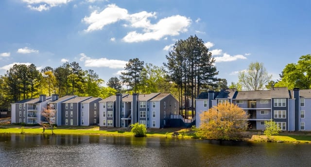 Apartment buildings overlooking a lake with trees and a blue sky.