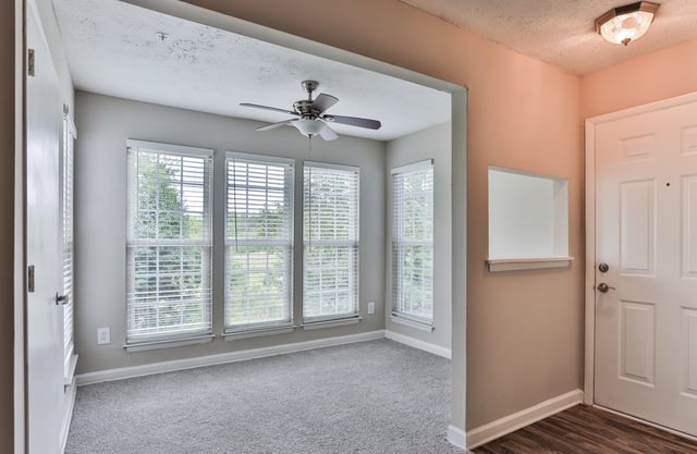 View of a living room with a ceiling fan and large windows, leading to an entryway with a white door.