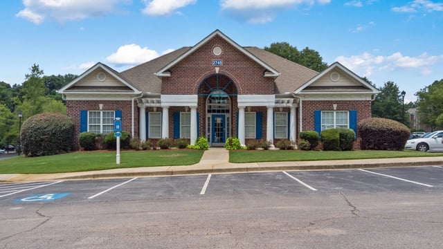Exterior of a brick building with blue shutters and white columns, featuring a parking lot with a handicapped space.