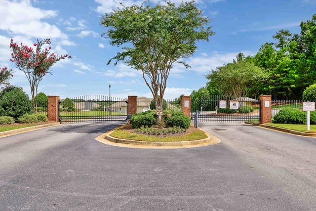 Gated entrance with brick pillars and black metal gates, surrounded by lush landscaping and trees.
