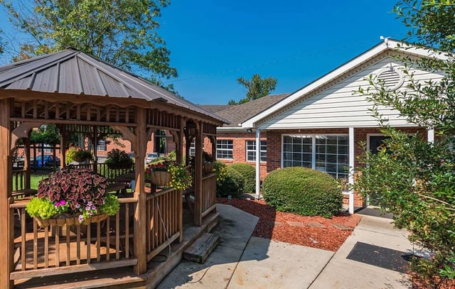 Wooden gazebo with hanging flower baskets next to a brick building with white siding.