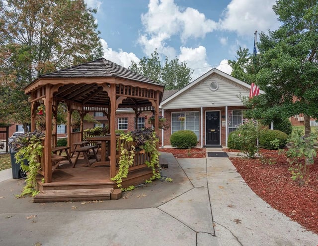 Wooden gazebo with picnic tables and a brick building with a black door.
