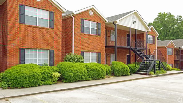 Exterior view of brick apartment buildings with green landscaping and parking lot.