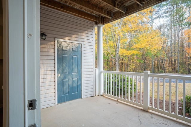 Exterior view of an apartment building entrance with a blue door and a white railing on a porch with autumn foliage in the background.