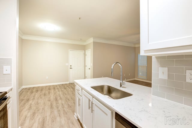 Modern kitchen with white countertops, stainless steel sink, and chrome faucet, opening into living area.