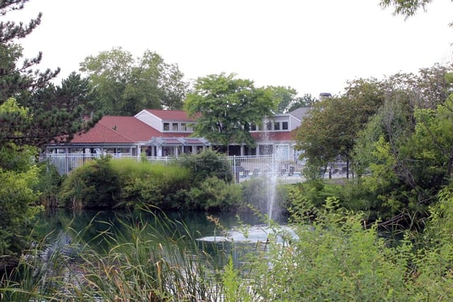A building with a red roof, surrounded by lush greenery, with a fountain in a pond in the foreground.