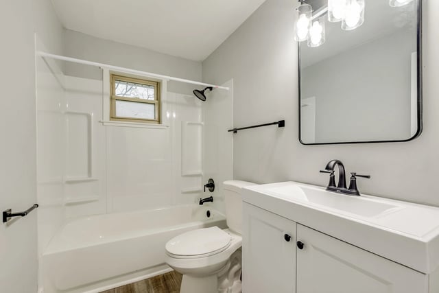 Clean and modern bathroom with a white vanity, sink, toilet, and a white bathtub/shower combo with a small window.