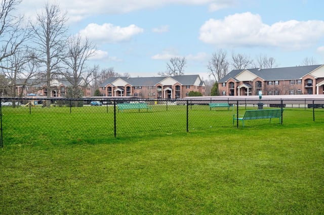 Fenced-in grassy dog park with benches and apartment buildings in the background.