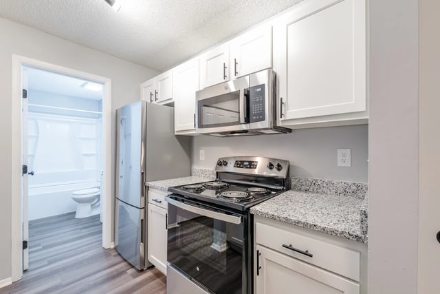 Modern kitchen with stainless steel appliances, granite countertops, and white cabinets. View into a bathroom with a bathtub and toilet.