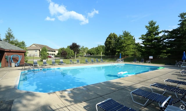 Sparkling outdoor swimming pool with lounge chairs and umbrellas, surrounded by lush greenery and apartment buildings.