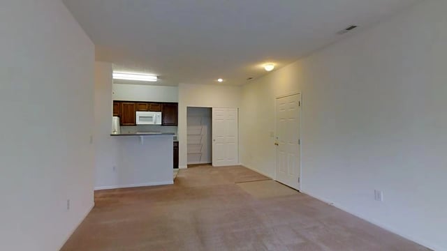 Living room and kitchen area with white walls and tan carpet. Features a breakfast bar, white appliances, and a closet.