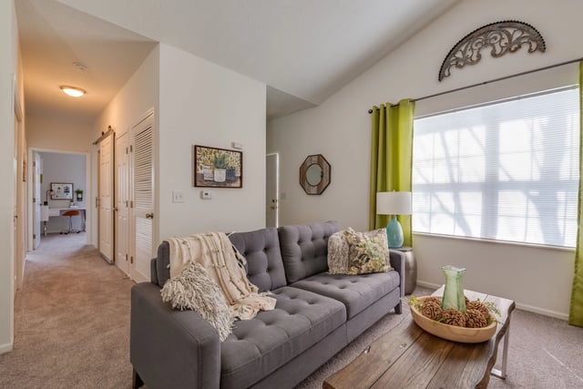 Gray tufted sofa with decorative pillows and throw blankets in a living room with a hallway and desk visible.