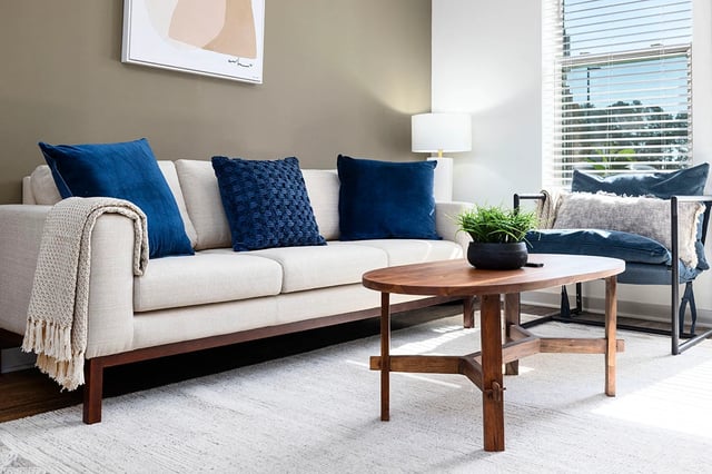 Modern living room with a cream sofa, blue accent pillows, a wooden coffee table, and a chair by the window.
