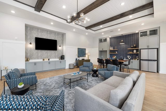 Modern community lounge area with seating, a large TV, and a dining area with a refrigerator.
