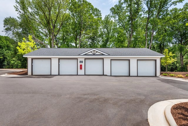 Six-car garage building with white doors and dark trim, set against a backdrop of lush green trees.