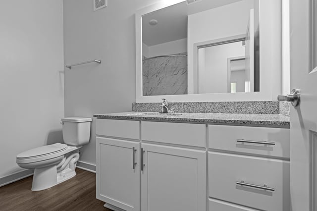 Modern bathroom with a granite countertop, white vanity, toilet, and a large mirror reflecting a tiled shower.