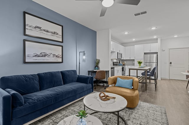 Living room and kitchen area of a modern apartment with a blue sofa, accent chair, and stainless steel appliances.