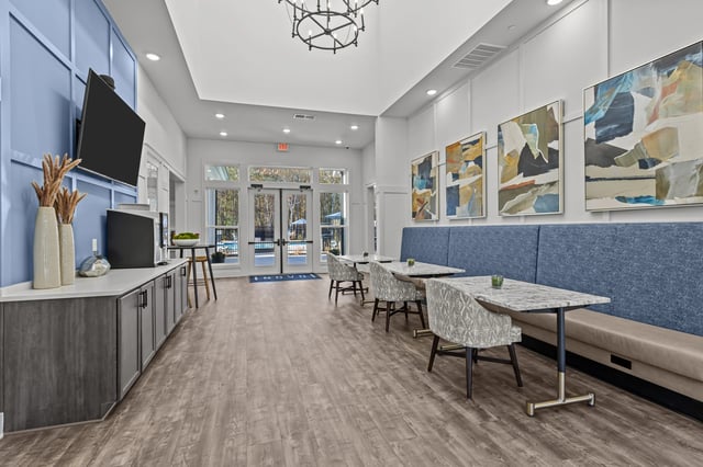 Seating area with tables and chairs in a community lounge with view of pool through glass doors.