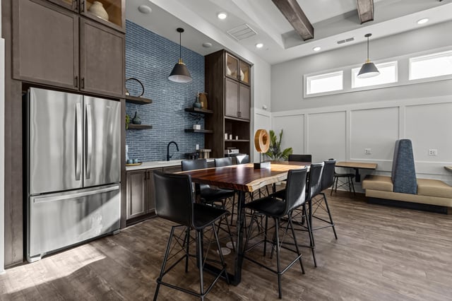 Community lounge area with a large live-edge dining table and seating, a full-size refrigerator, and a stylish blue brick backsplash.