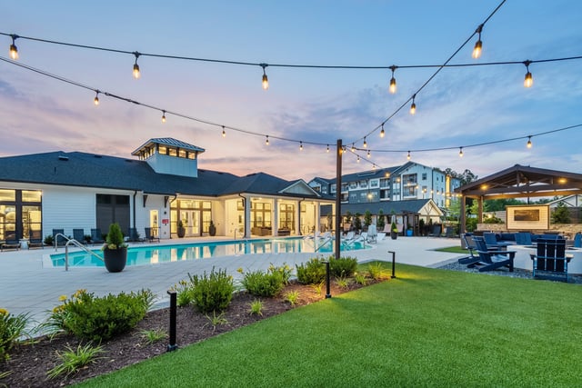Resort-style pool area with lounge chairs, fire pit, and outdoor TV, illuminated by string lights at dusk.