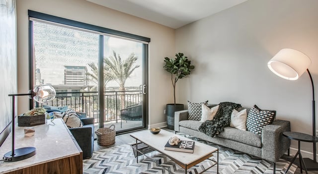 Modern living room with a grey sofa, marble coffee table, and large window overlooking a cityscape.