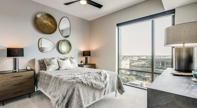Modern bedroom with a king-size bed, plush bedding, and city views from a large window.