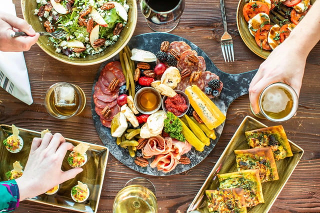 A table full of food, including a charcuterie board, salad, deviled eggs, and pizza slices, with hands reaching for the food and drinks.