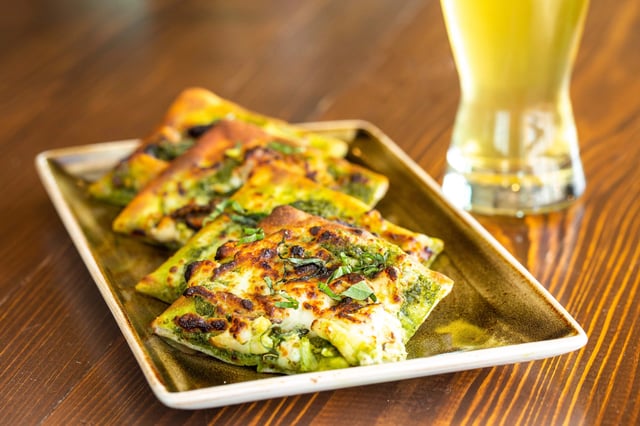 Flatbread with pesto and cheese served on a platter with a glass of beer.