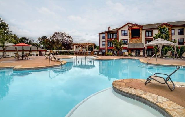 Resort-style swimming pool with lounge chairs and cabanas, apartment building in the background.