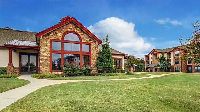 Exterior of apartment building with stone accents and large windows, featuring a grassy courtyard and volleyball net.