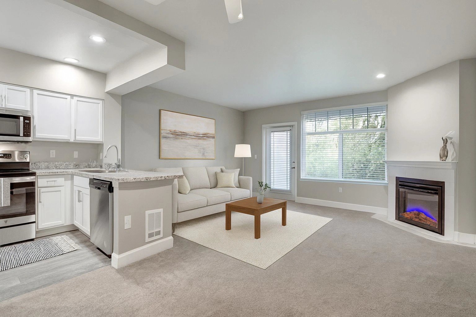 Open-concept living area with a sofa, coffee table, fireplace, large window, and adjacent kitchen.
