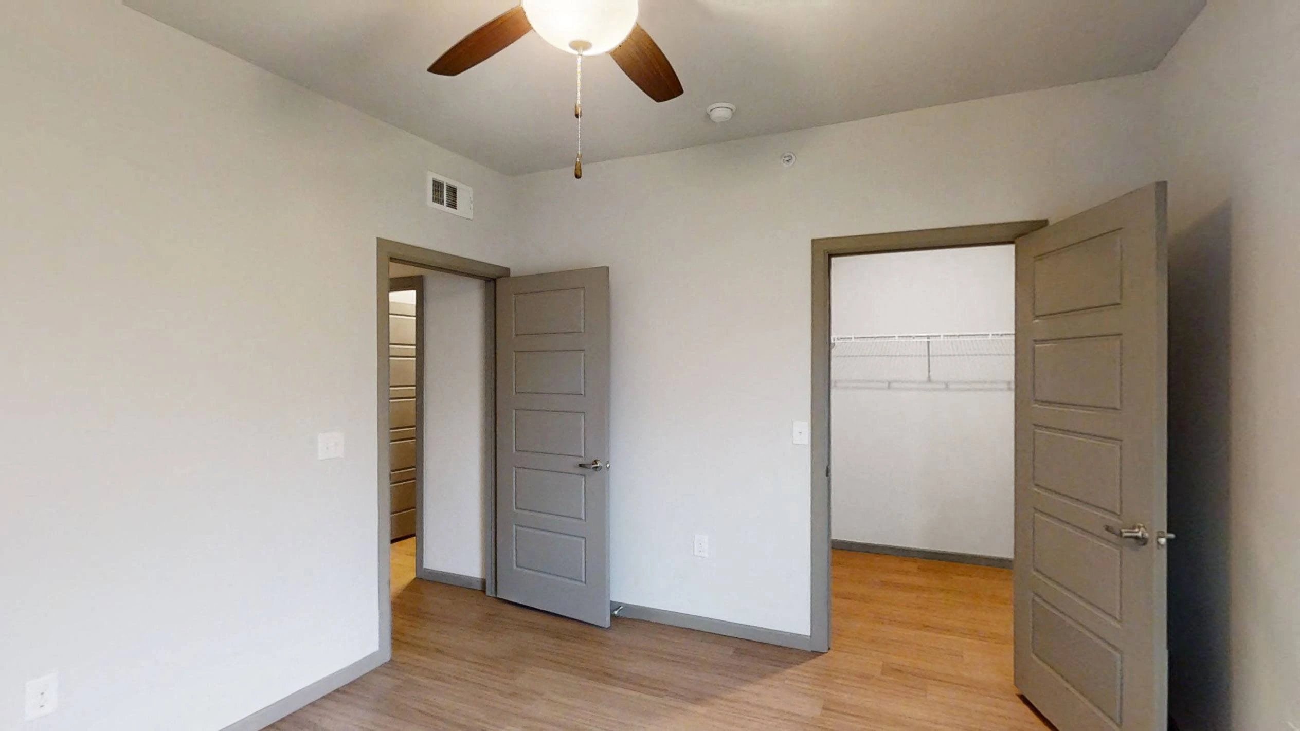 Bedroom interior with ceiling fan, neutral walls, wood flooring, and closet shelving.