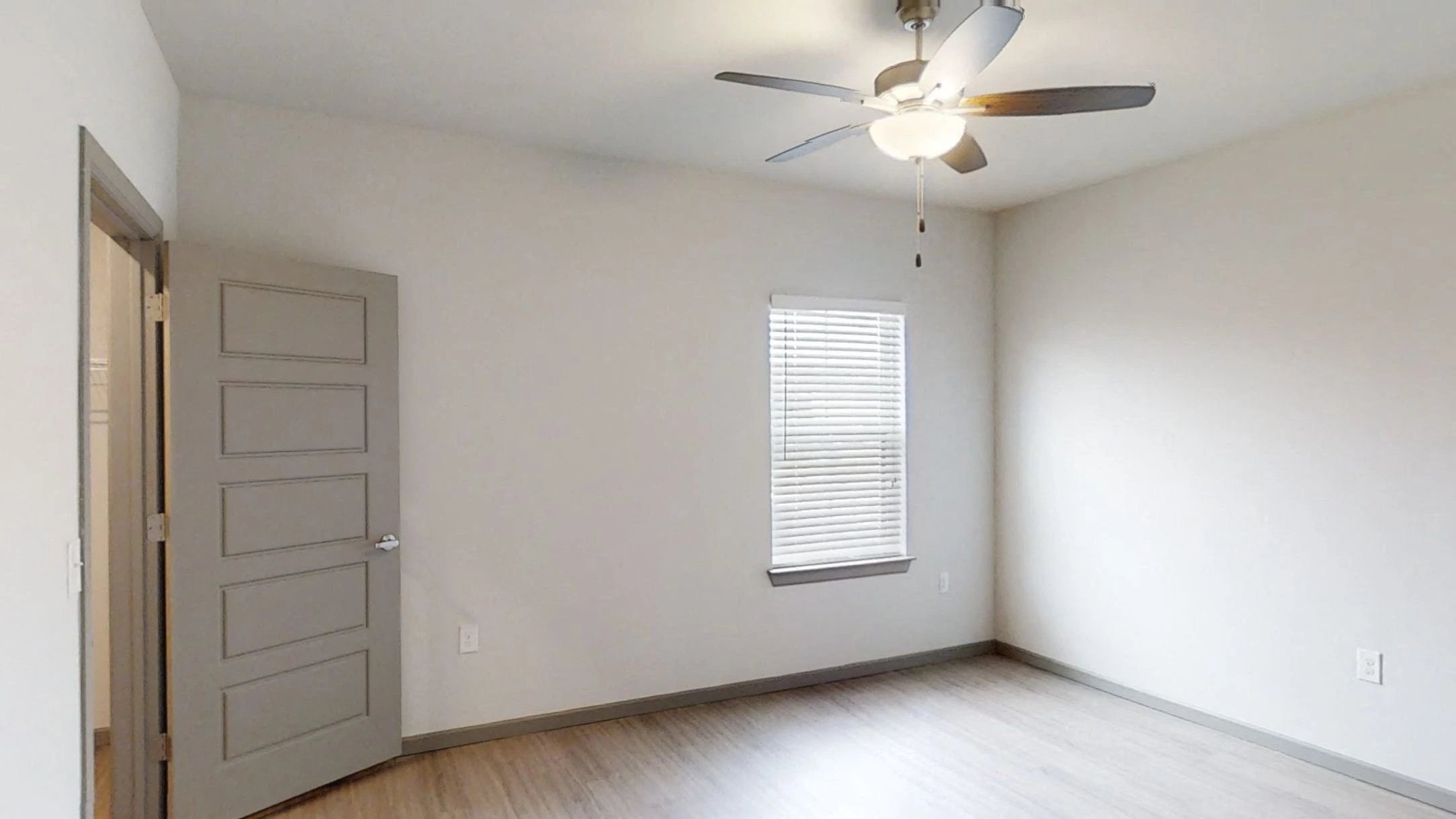 Empty apartment room with a ceiling fan, doorway, and a window with blinds.