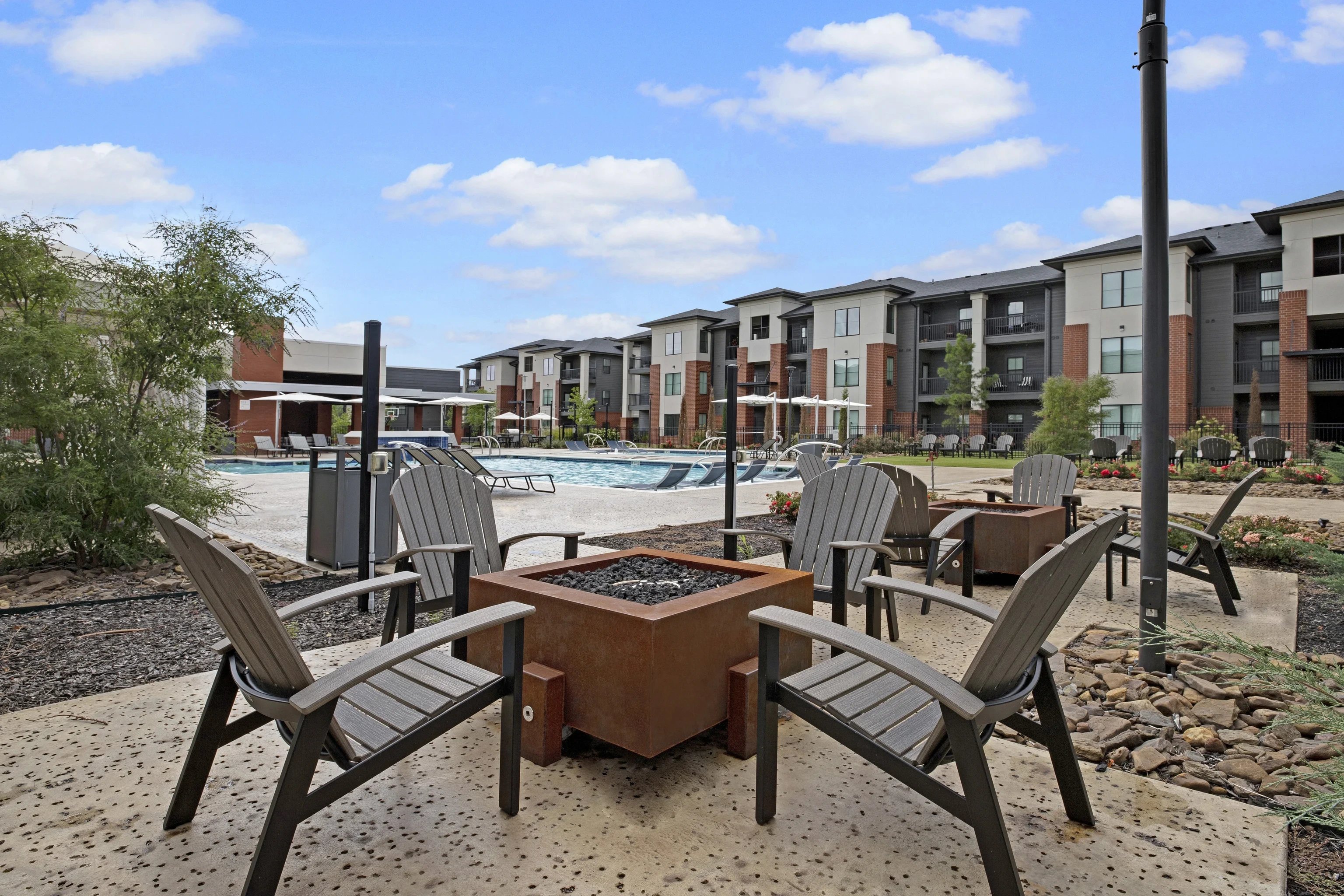 Outdoor community pool area with lounge chairs and a central fire pit near apartment buildings.