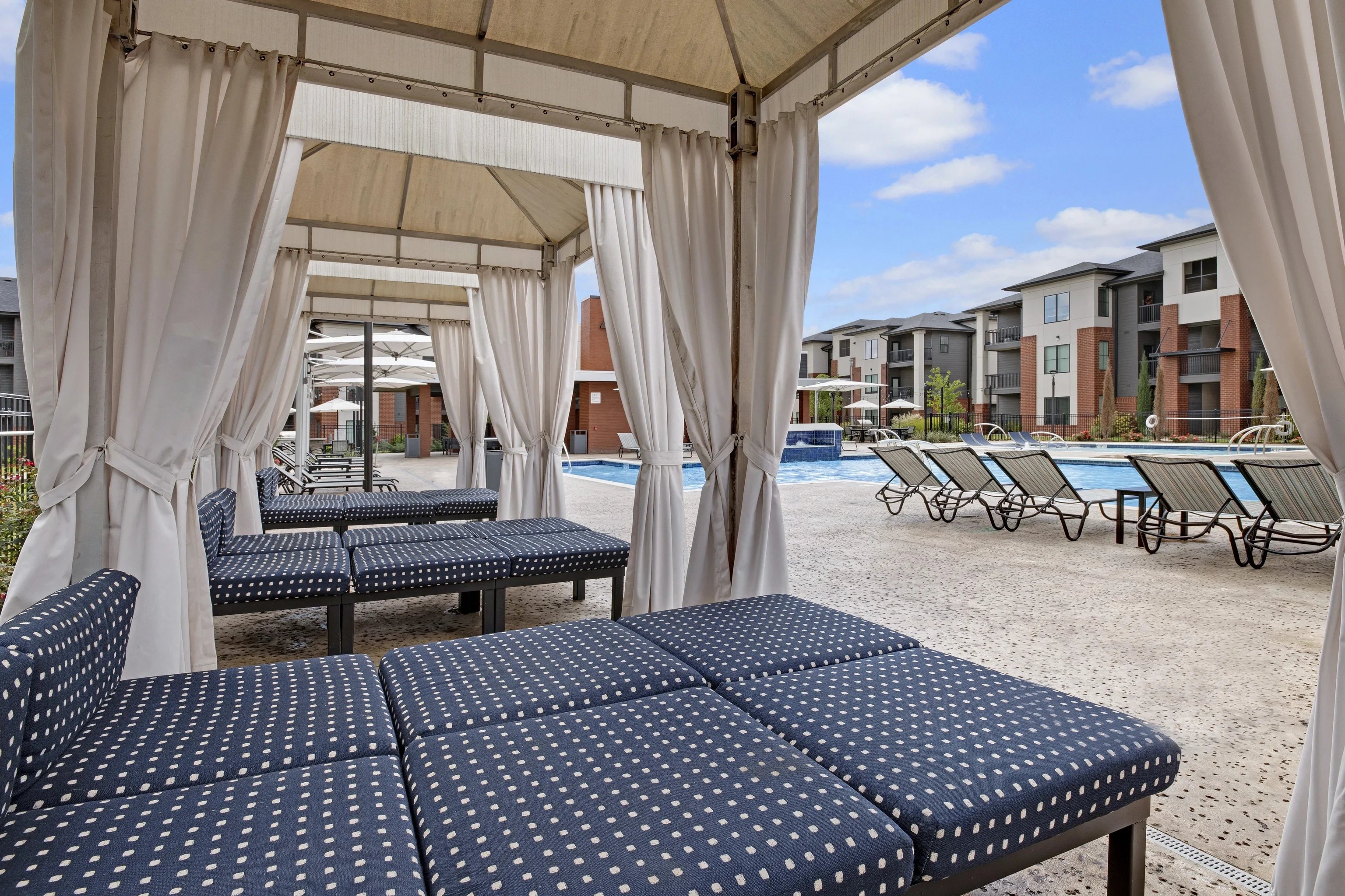 Pool area with white cabanas, flowing curtains, and rows of blue-dotted lounge chairs.