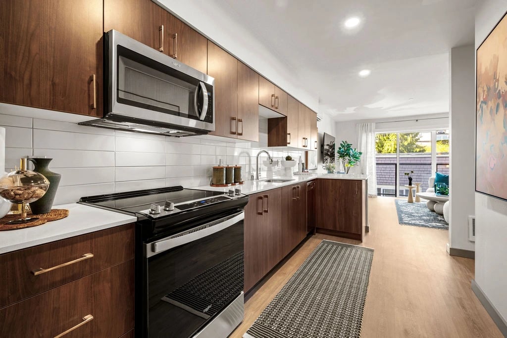 Modern apartment kitchen with dark wood cabinets, stainless appliances, white subway backsplash, and living area beyond.