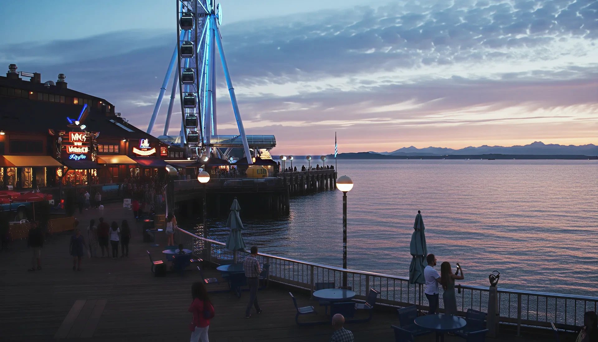 Sunset over a waterfront boardwalk with a lit Ferris wheel, restaurant lights, and people by the railing.